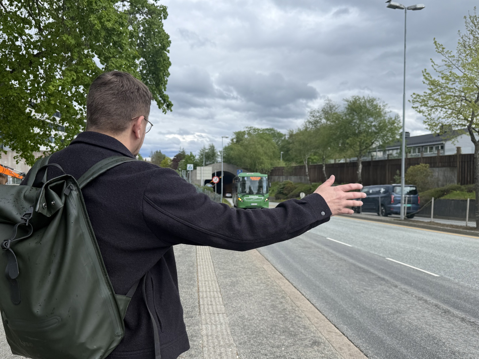 Mann med sekk rekker ut hånden for å stoppe buss på bussholdeplass.