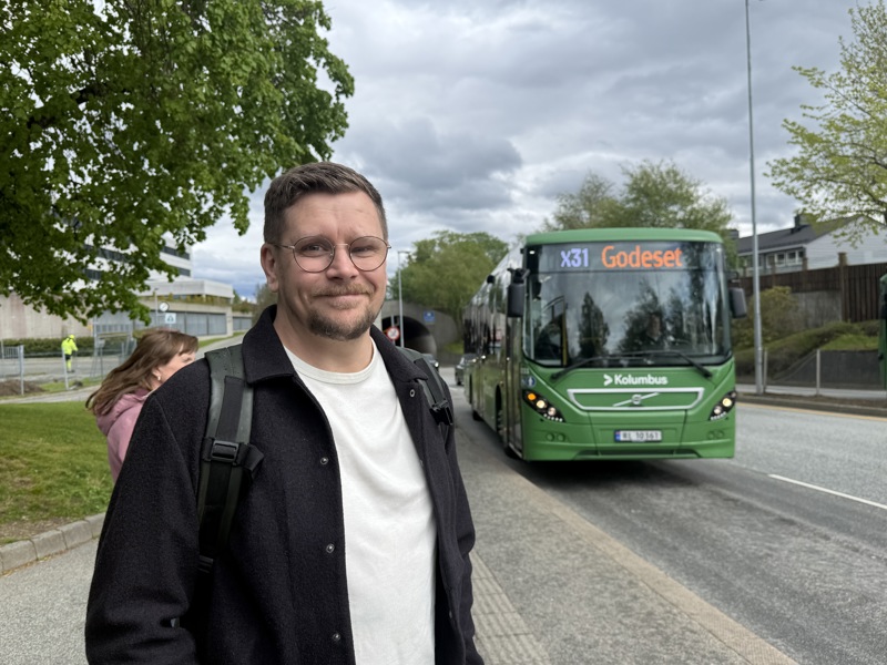 Mann med sekk står på bussholdeplass og smiler. En grønn buss kommer i mot holdeplassen i bakgrunnen.