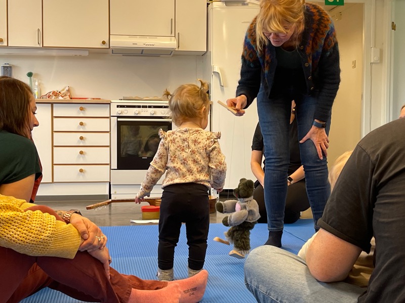 Inne i Veden åpen barnehage. Liten jente ser på musikkpedagog som hoder en dukke. Den lille jenta står på en blå matte. 