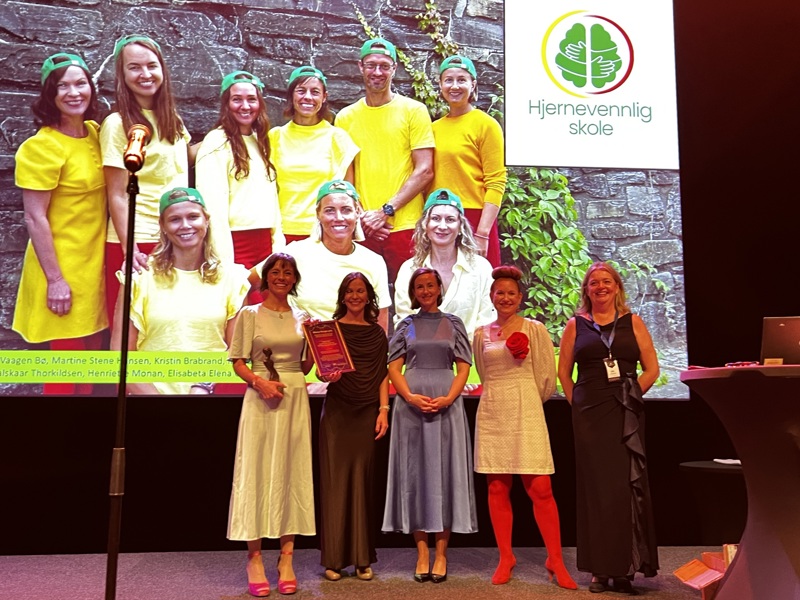 De tre prismottakerne fra Stavanger står sammen med to prisutdelere fra Mensa Norge i fine kjoler foran et gigantisk bilde av hele hjernevennlig-teamet i skolehelsetjenesten i Stavanger.