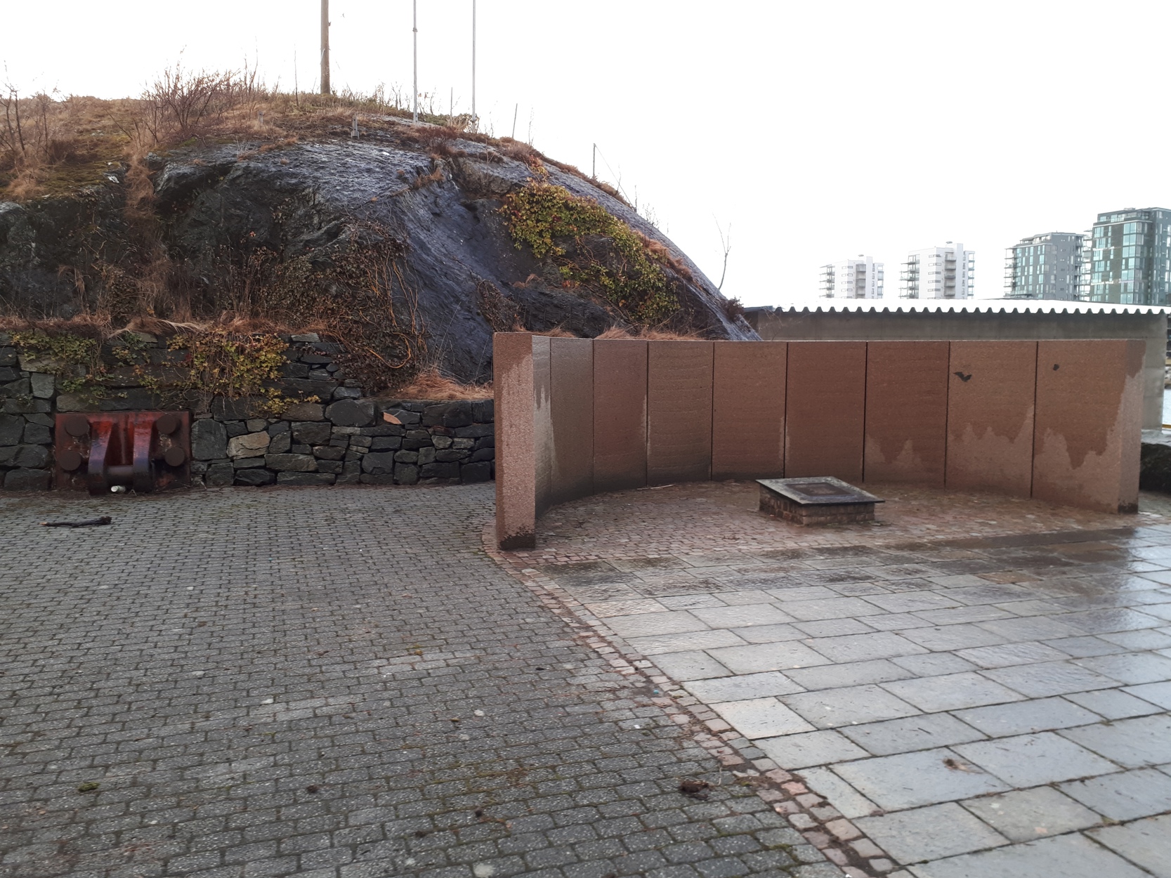 Monument over Concem-ulykken en regnværsdag, med boligblokker i bakgrunnen.