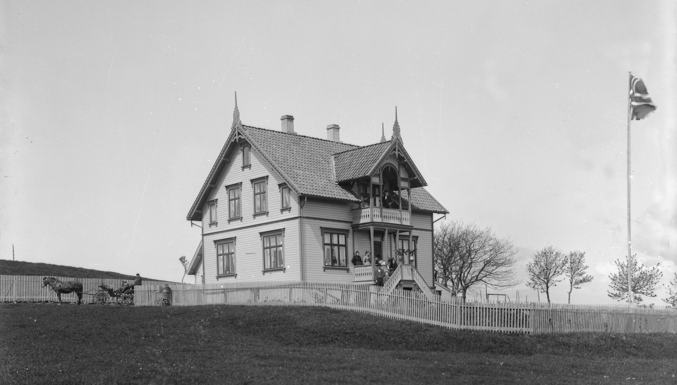 Villa i sveitserstil, med stakittgjerde, hage med trær, flaggstang med norsk flagg, og hest med vogn utenfor