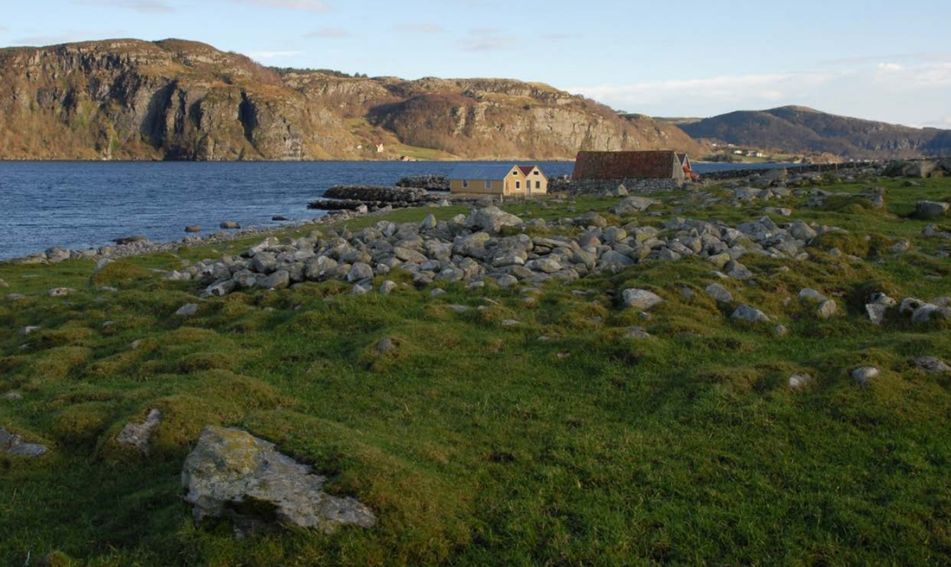 Beitemark med gravrøyser i forgrunnen, naust, moloer og steingarder, mot blå fjord og bratt landskap over fjorden. Blek himmel.