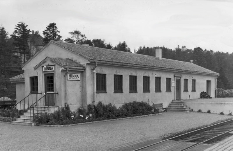 Et svart-hvittbilde av en enkel, enetasjes, murt stasjonsbygning i funkisstil med påskrift HINNA over døra.