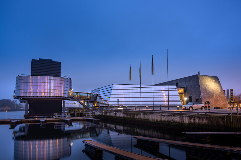 Norsk Oljemuseum sett fra  Blå Promenade ved Børevika, fotografert i skumringslys en kveld med klarvær. Fjorden ligger blikkstille, og speiler oljemuseet, som igjen speiler byens lys i den  nærmeste sylinderplattformen som står i sjøen.