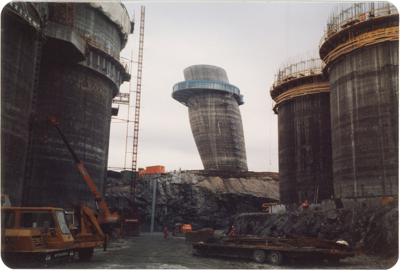 Plattformbyggevirksomhet: Fire store plattformelementer støpes  bit for bit i høyden. Armeringsjern stikker opp over betongen. Betongen, bakken og fjellet som er skåret bort i store stykker er alt grått, imens forskalingene, kraner og anleggsmaskiner, konteinere og anleggsarbeiderne skiller seg ut i oransje. Sentralt i bildet, noe bak byggeaktiviteten, står Skråtårnet, med sin blåfargede, utkragede etasje.