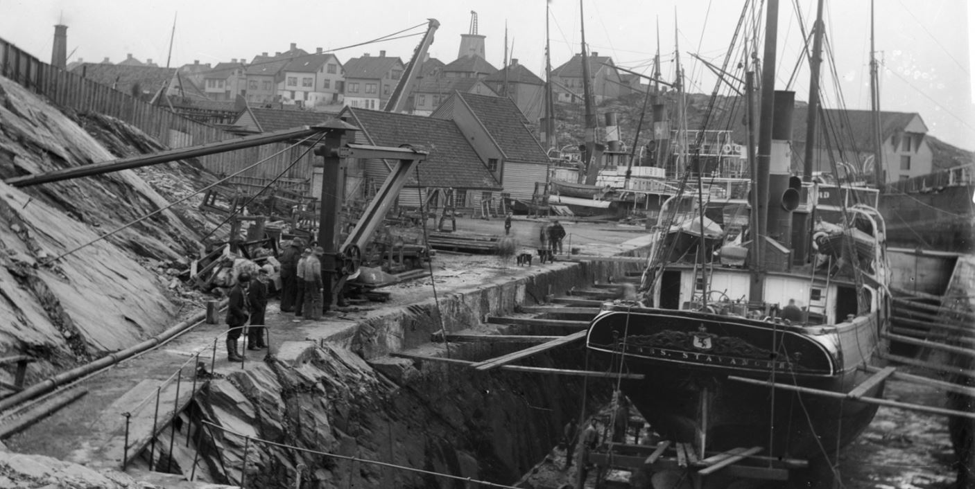 Et gammelt bilde av et dampskip i tørrdokk. To velkledde menn kikker ned i dokken, og arbeidere ses både nede ved skroget og oppe ved kranen over. I bakgrunnen ses boligstrøk, et sjøhus, flere skip og en vindmølle øverst.