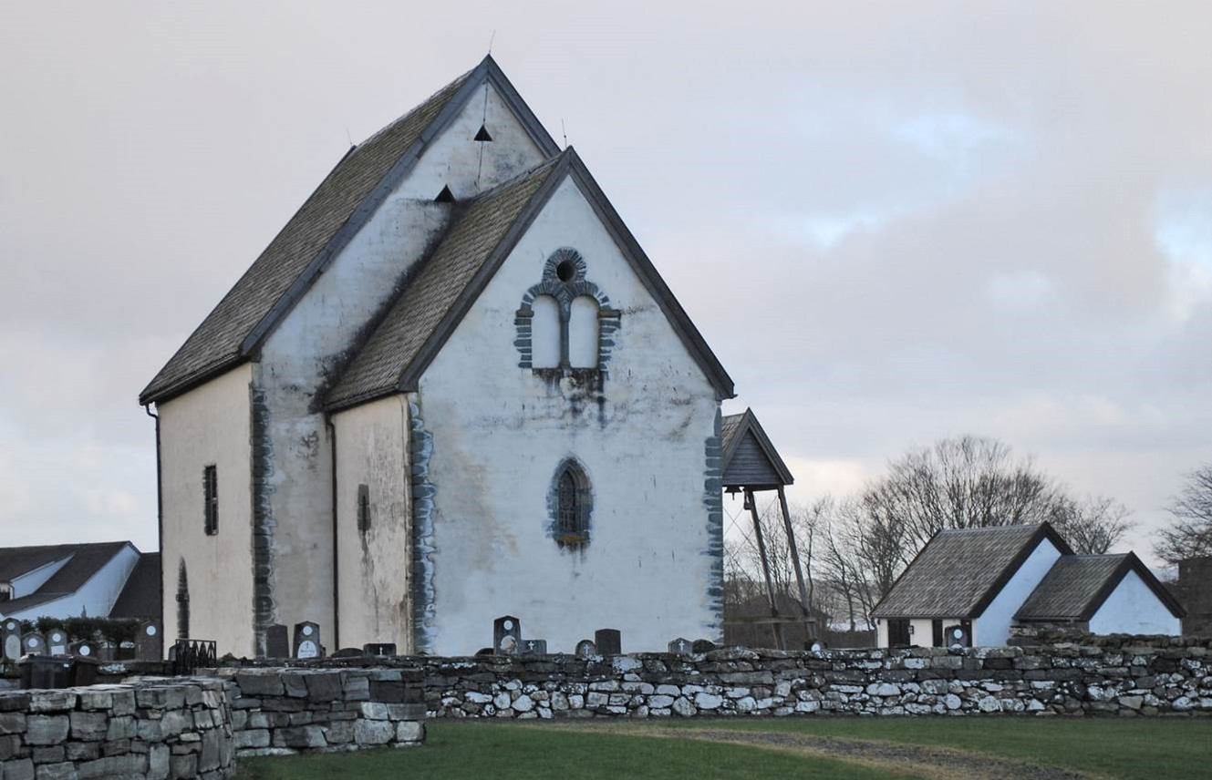 Sørbø kirke med litt av kirkegården og kirkegårdsmuren foran. Bildet ser ut til å vær tatt om vinteren; det er ikke løv på trærne, og himmelen er mørk til å være fotografert om dagen.