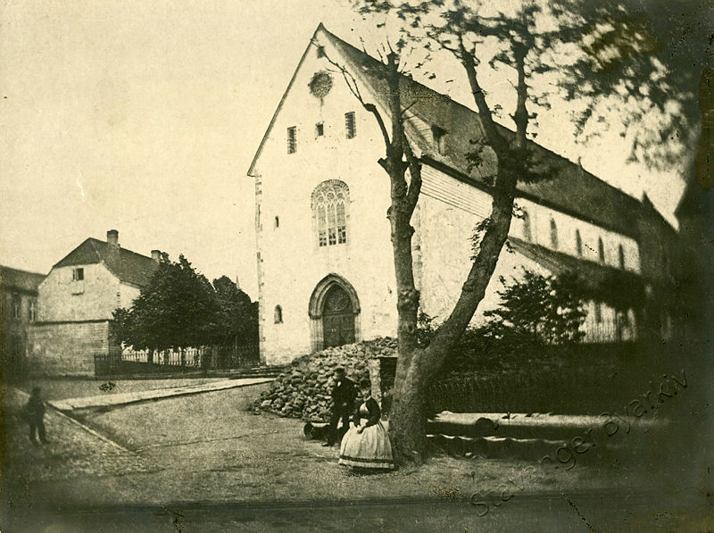 Eit gamalt, svart-kvitbilete av Stavanger Domkirke, litt av Domkirkeplassen og muren kring Kongsgård. Det er tre menneske i biletet: Ein mann eller gut? lengst mot venstre biletkant, og ein mann og ei dame i stor kjole midt føre kyrkja. Det er nokre tre nær dei to, og ei trerekke mellom Domkirken og Rådstuen