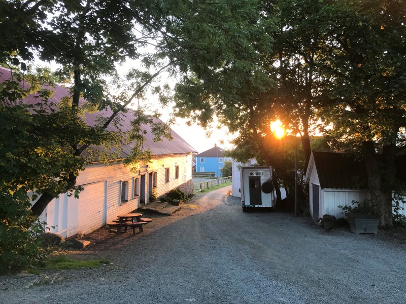 Et bilde av en stall og et våningshus i en bakke. Soloppgangen farger bildet rødlig. Det står en hestetransporthenger i tunet.