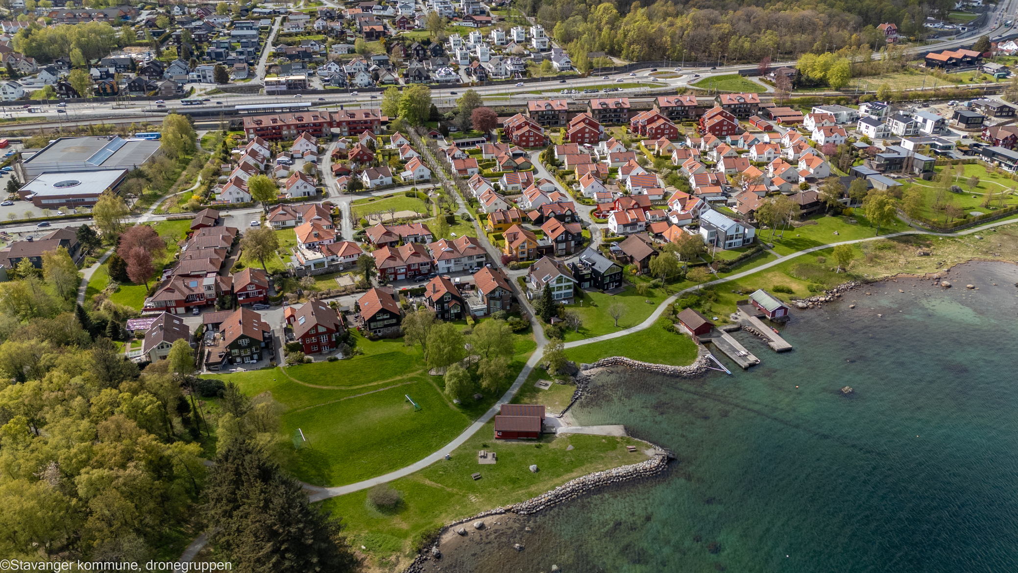 Dronebilde som viser boligfeltets plassering ved Gandsfjorden. Vi ser alléer, ulike boligtyper, båthus, strand, badebrygge og tursti.