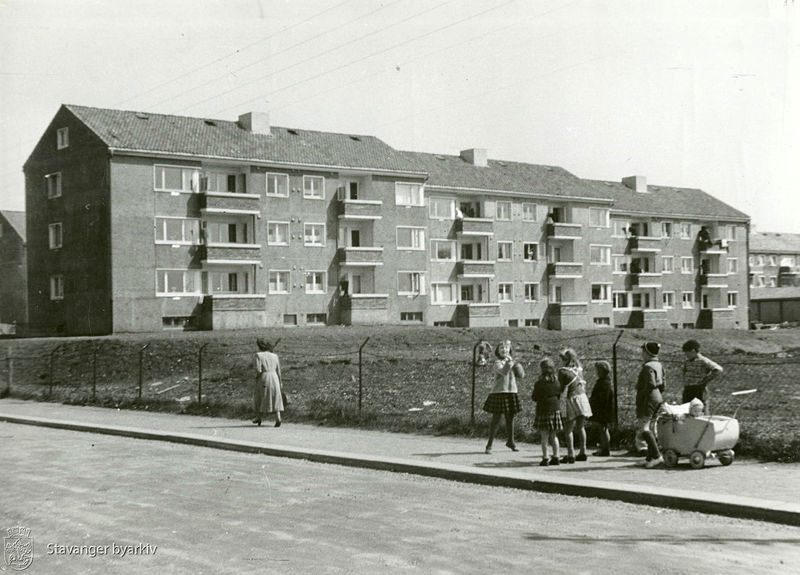 Svart/hvitt-bilde av barn som leker med bl.a. dukkevogn i forgrunnen. Boligblokker i bakgrunnen. 