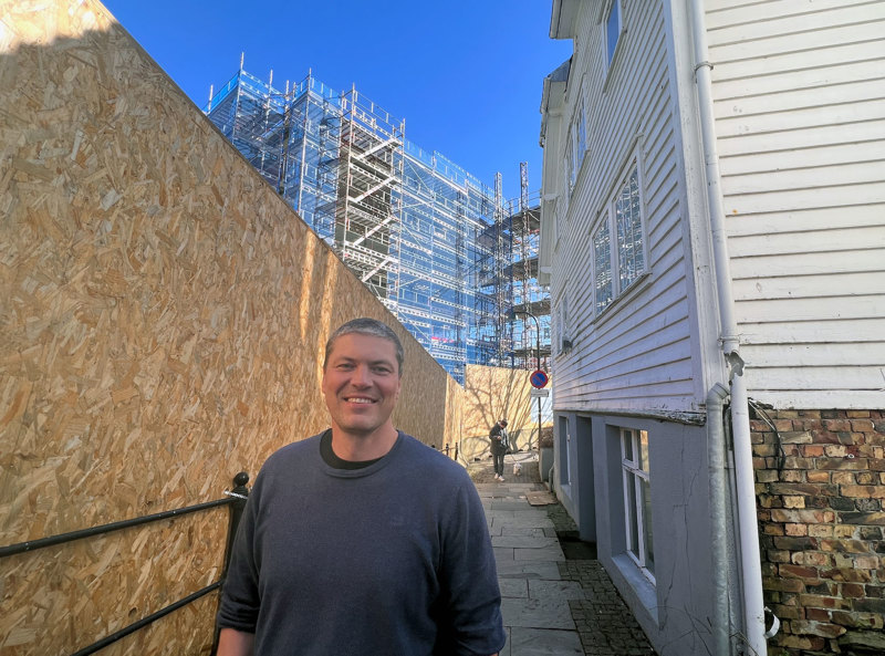 Byggesaksjef Sven Norland står mellom et byggegjerde og et gammelt trehus, med et byggeprosjekt i bakgrunnen. 