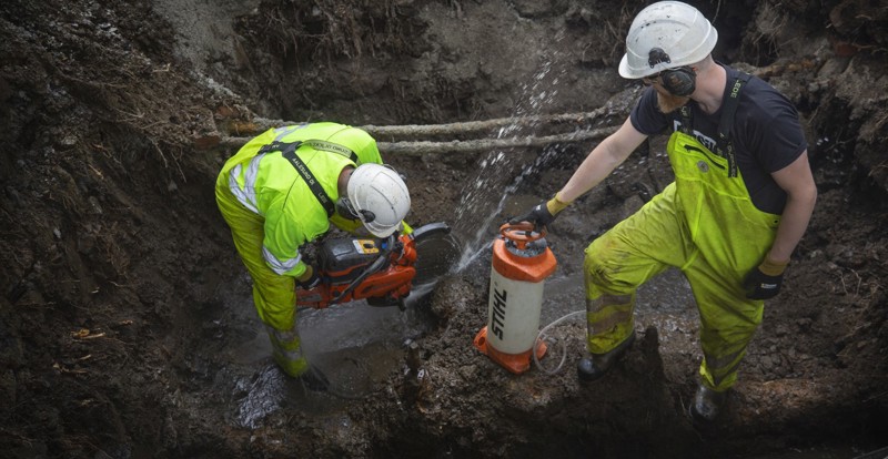 To menn i hjelp og arbeidstøy står i en dyp grøft og kutter gamle avløpsrør. Foto: Elisabeth Tønnessen.