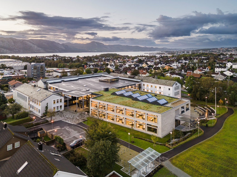 Bilde av nybygget ute, med de eldre byggene rundt. Byggets arkitektur tar vare på alle de ulike møtene som er gjennom skoledagen, i undervisningssituasjon, småprat i korridorene og aktivitet utendørs i friminuttene. 