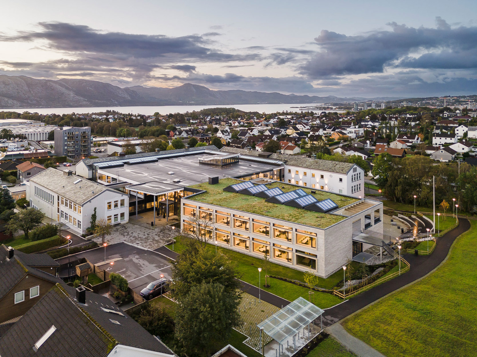 Bilde av nybygget ute, med de eldre byggene rundt. Byggets arkitektur tar vare på alle de ulike møtene som er gjennom skoledagen, i undervisningssituasjon, småprat i korridorene og aktivitet utendørs i friminuttene.