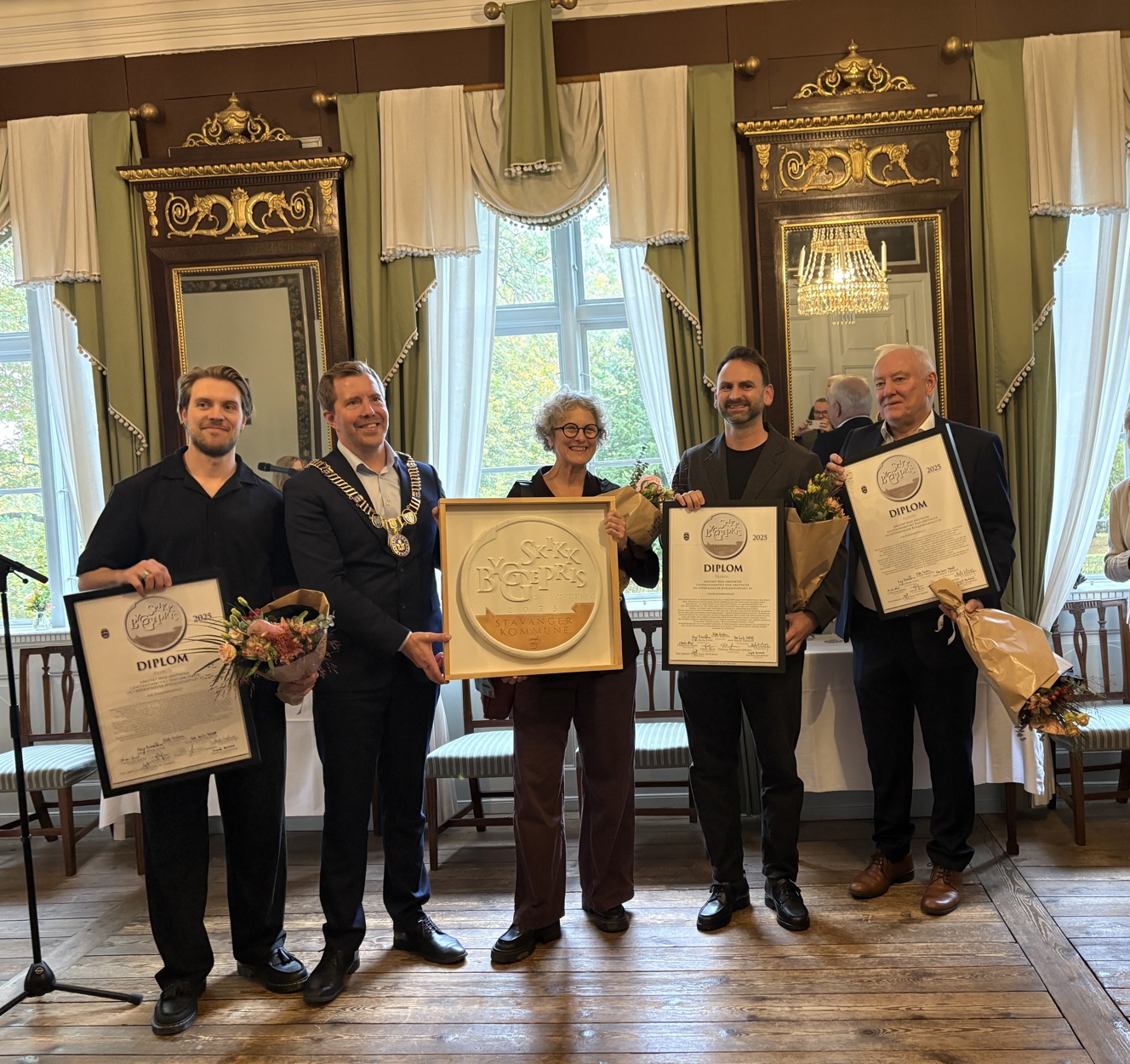 Fire menn og en kvinne i staselige omgivelser, med diplomer og plakett