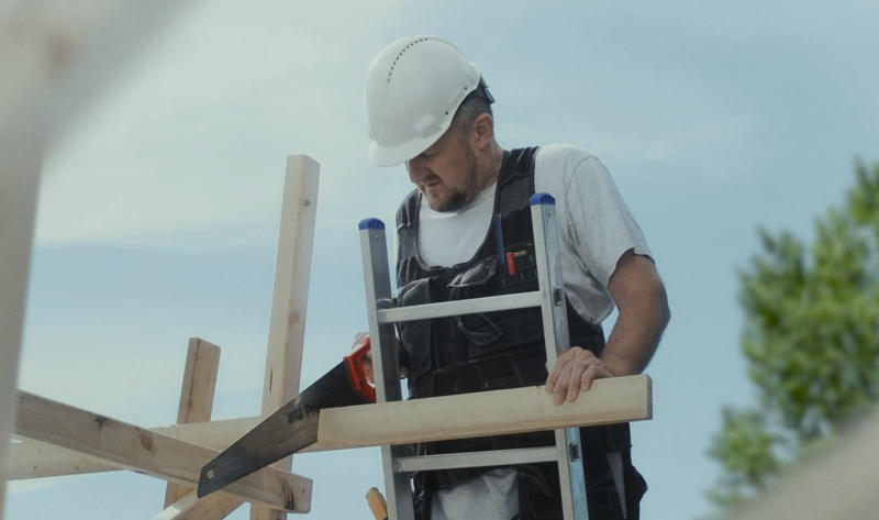 Snekker som står og sager av bjelken han støtter stigen sin på. Skjermbilde fra promoteringsfilm for Tettpå-kampanjen 2022