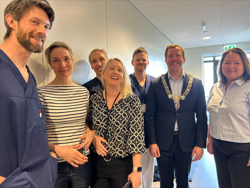En blid bukett leger, administrativt ansatte og ordfører står samlet i gangen på nye Tjensvoll legekontor. Tre av legene har blå kitler på. Ordføreren har ordførerkjede.