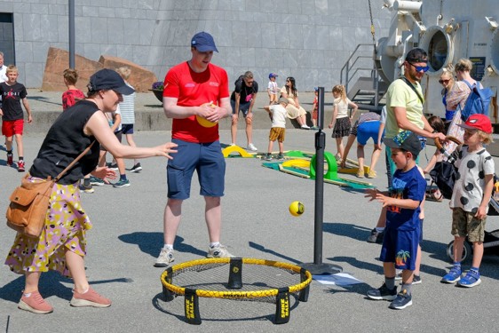 Vi ser en rekke mennesker som står på en stor plass og holder på med ulike aktiviteter. Fremst står en kvinne som kaster en ball i en rundt nett som ligger litt over bakken. To barn følger spent med når damen kaster ballen.