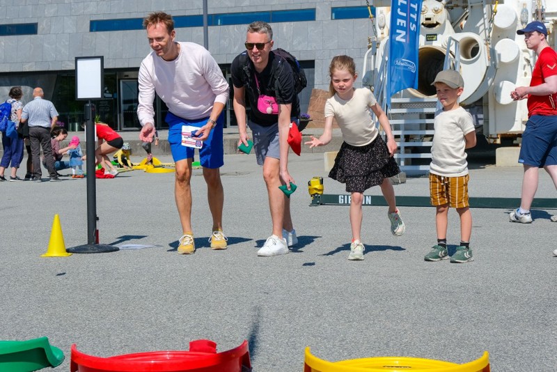 Vi ser to menn og to barn som står på en rekke. De kaster ball, og prøver å treffe et hull i en konkurranse.
