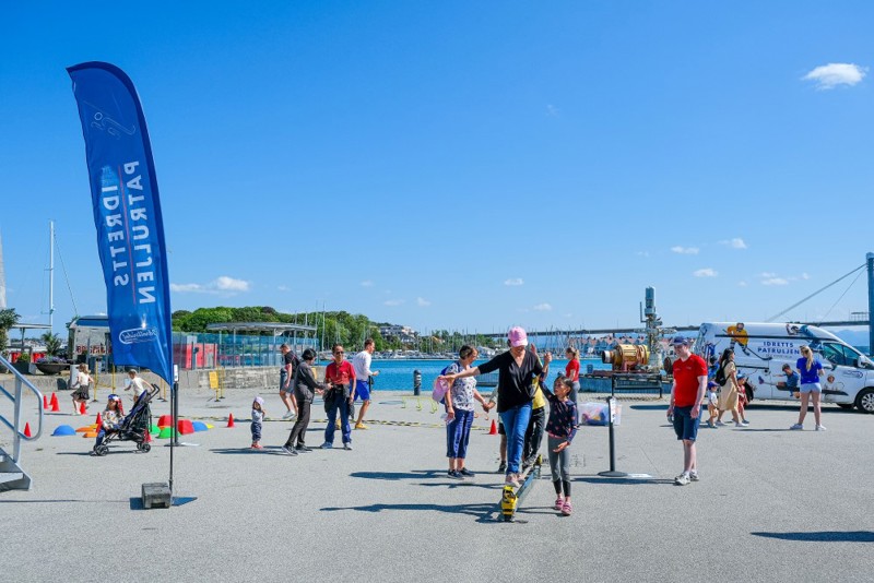 Vi ser flere mennesker som gjør aktiviteter på en stor uteplass. En dame i midten av bildet forsøker å balansere på en kant.