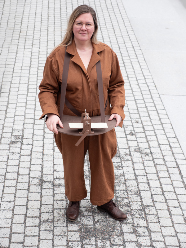 Kunstneren Anne Marte Overaa iført en brun buksedress, ser inn i kameraet. Hun har en liten grafisk presse rundt halsen.