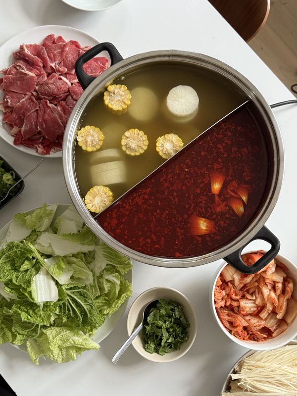 Et dekket bord sett ovenfra. På en hvit duk står en hotpot-gryte med to versjoner av kraft, den ene er klar og den andre er dyp rød. Et fat med oppskåret storfekjøtt, et fat med salatblader, en skål med koriander og en skål med kimchi står rundt gryten.