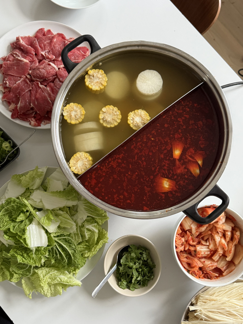 Et dekket bord sett ovenfra. På en hvit duk står en hotpot-gryte med to versjoner av kraft, den ene er klar og den andre er dyp rød. Et fat med oppskåret storfekjøtt, et fat med salatblader, en skål med koriander og en skål med kimchi står rundt gryten.