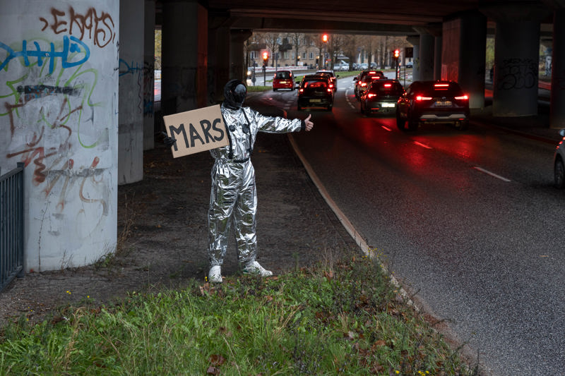 En person ikledd en sølvfarget dress og sort maske står langs en vei og haiker. På skiltet han har med seg, som viser bilistene destinasjonen han ønsker å reise til, står det MARS.