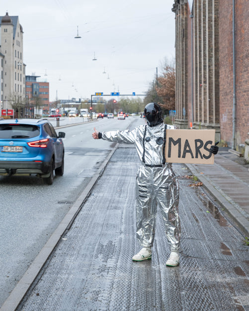 En person ikledd en sølvfarget dress og sort maske står langs en vei og haiker. På skiltet han har med seg, som viser bilistene destinasjonen han ønsker å reise til, står det MARS.