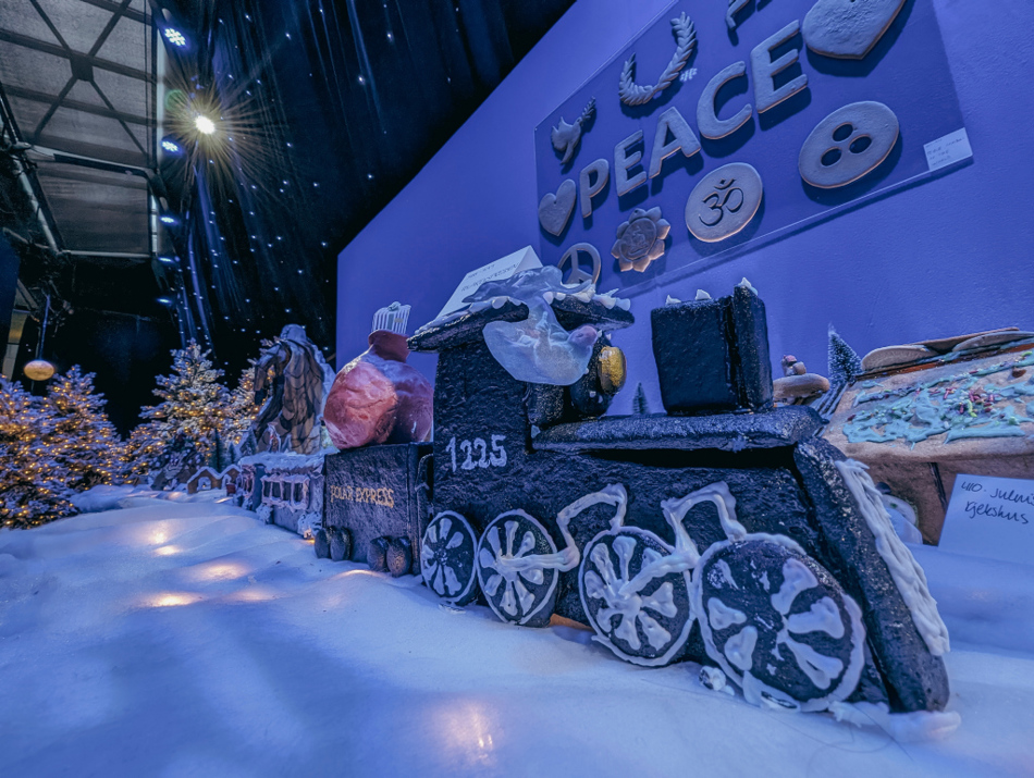 Et stort pepperkaketog med detaljerte hjul og lokomotiv står på et snødekket underlag. Toget er dekorert med hvit glasur og har nummeret «1225» på siden. I bakgrunnen er det flere pepperkakefigurer og juletrær med lys. På veggen bak toget henger ordet «PEACE» sammen med symboler og dekorasjoner i ulike former.