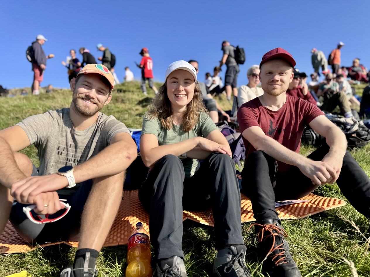 En kvinne sitter mellom to menn, alle smiler. Bak dem er turkledde mennesker.