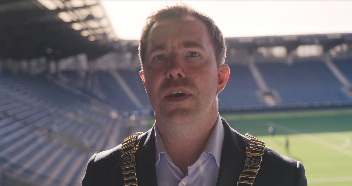 Ordfører Tormod W. Losnedal står på stadion med banen i ryggen og ser inn i kamera. Han synger.