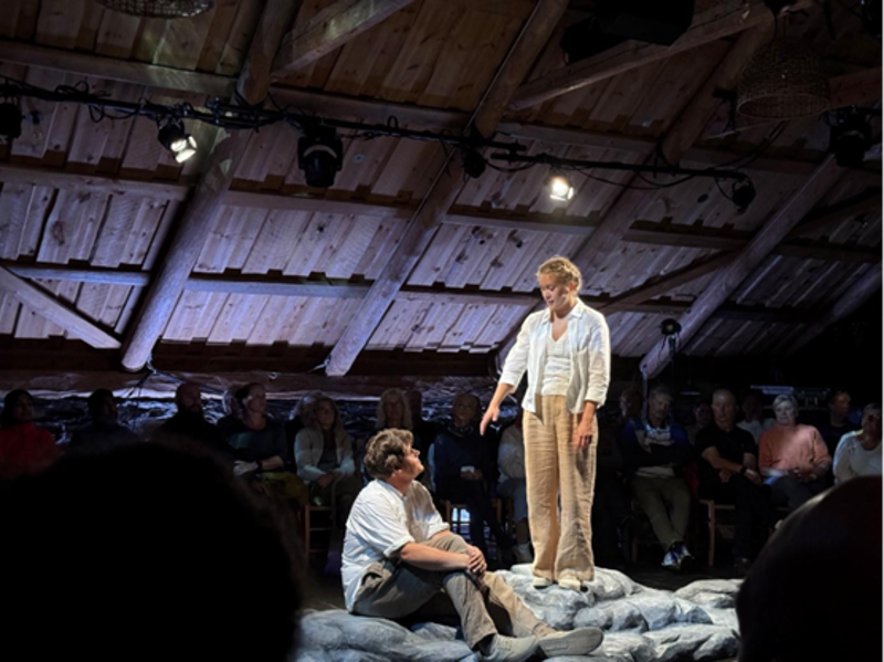 Bilde fra forestillignen. En kvinne strekker ut armen til en mann som sitter. 