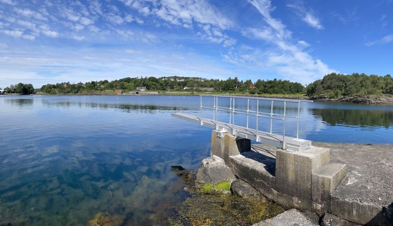Stupebrett i stål på betongfundament. Himmel med hvite skyer speiler seg i mørkeblå sjø.