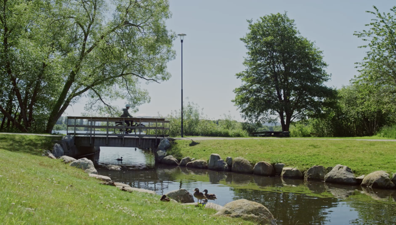 Bilde av bekk ned til Mosvatnet. Syklist på brua i bakgrunnen. Fin sommerdag.