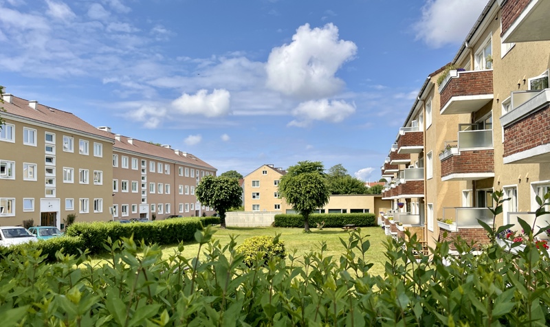 Vi ser bildet av lave blokker på Misjonsmarken i Stavanger. Med små balkonger ut mot en hage på begge siden, og med et murhus i midten. Det er grønt og frodig med grønne hekker og trær.
