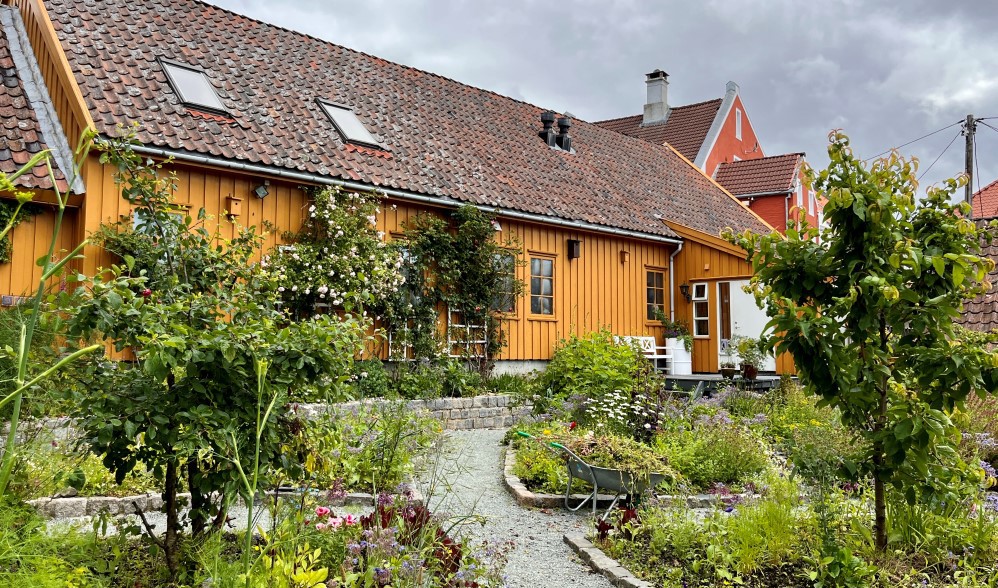 Vi ser det gamle okergule huset inne i gården, med frodig inndelt hage, med masse blomster og nyttevekster.