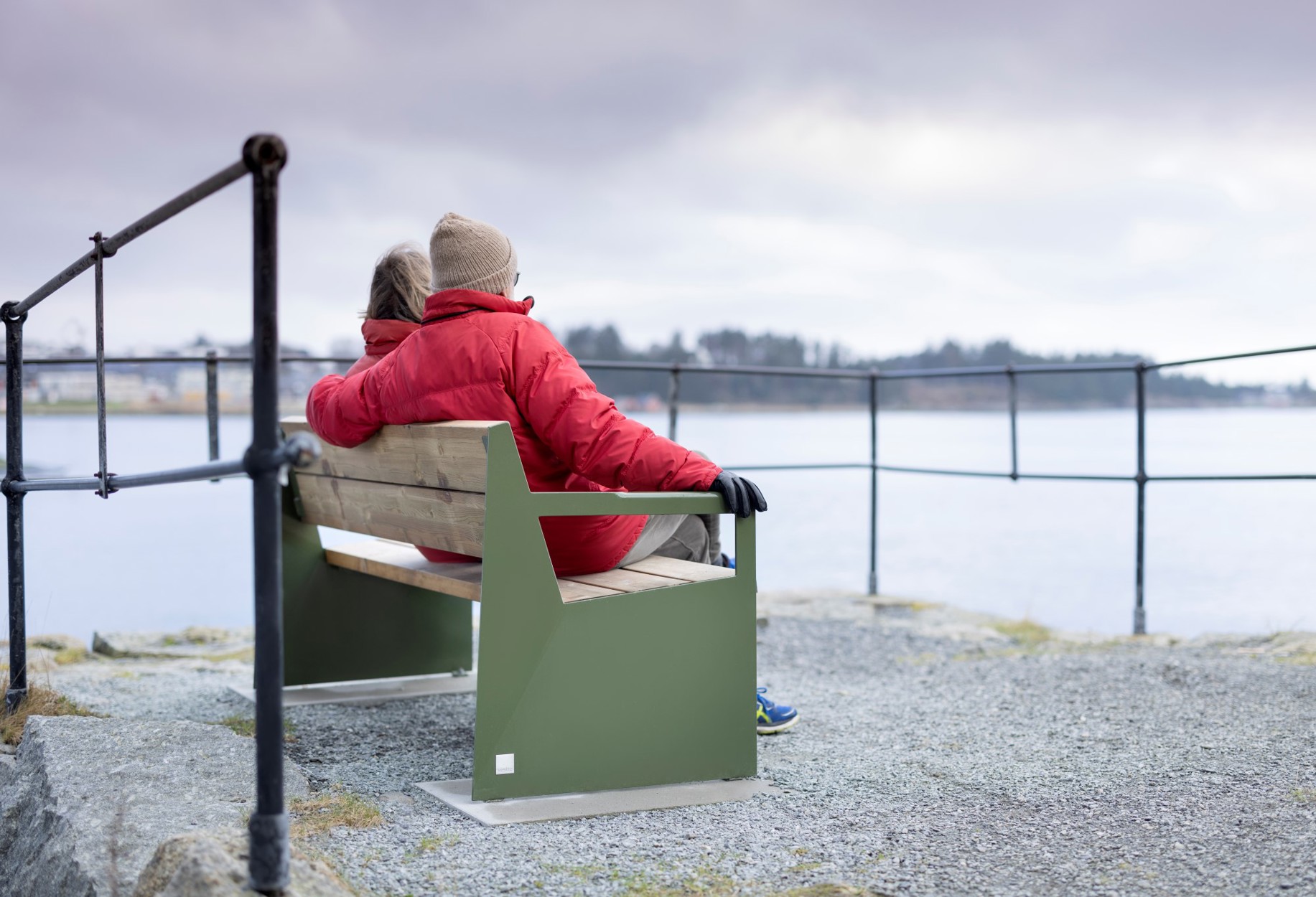 Vi ser et eldre par som sitter på en grønn benk ved sjøen.