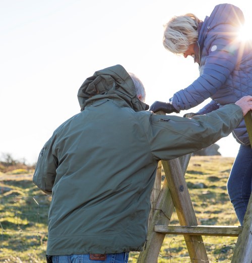 Vi ser to personer som skal gå over en gjerdestolpe i tre. En mann hjelper en dame over.