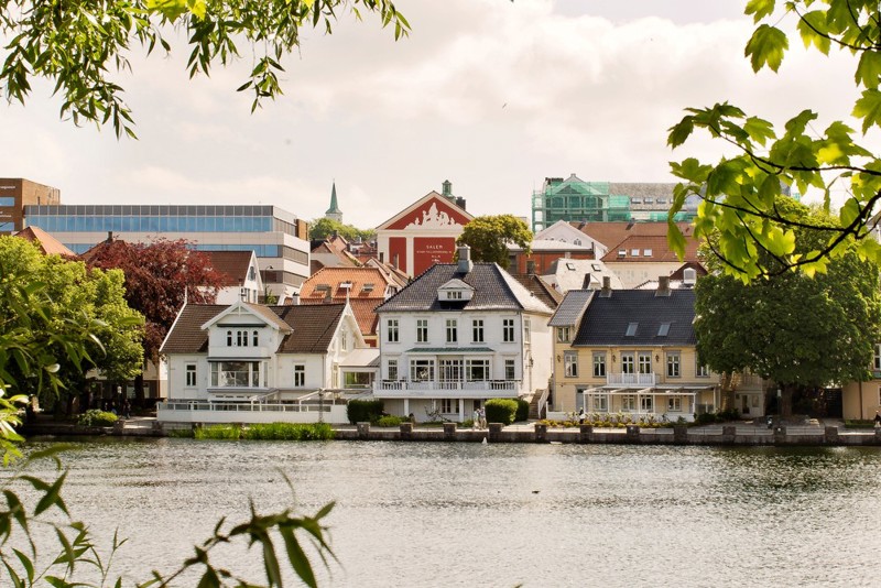 Bildet viser et utsnitt av utsikten ved Breiavatnet. Vi ser flere hus som ligger ved vannet. Det er sommer, grønt og folk går forbi. 