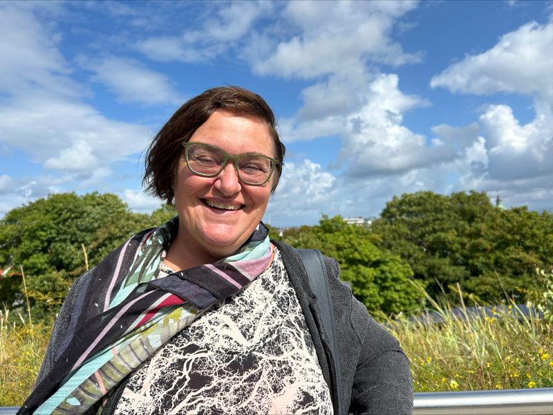 Portrettbilde av May Britt Jensen. Hun har mørkt krøllet hår, briller og et bredt smil. I bakgrunnen er den grønne trær, og solen skinner.