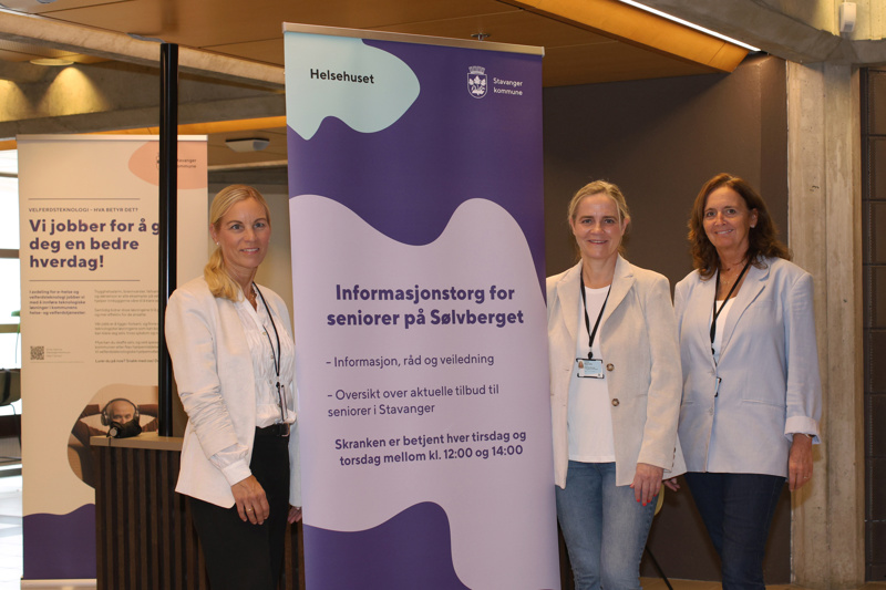Tre damer står med en blå roll up med informasjon om seniortoget på mellom seg.