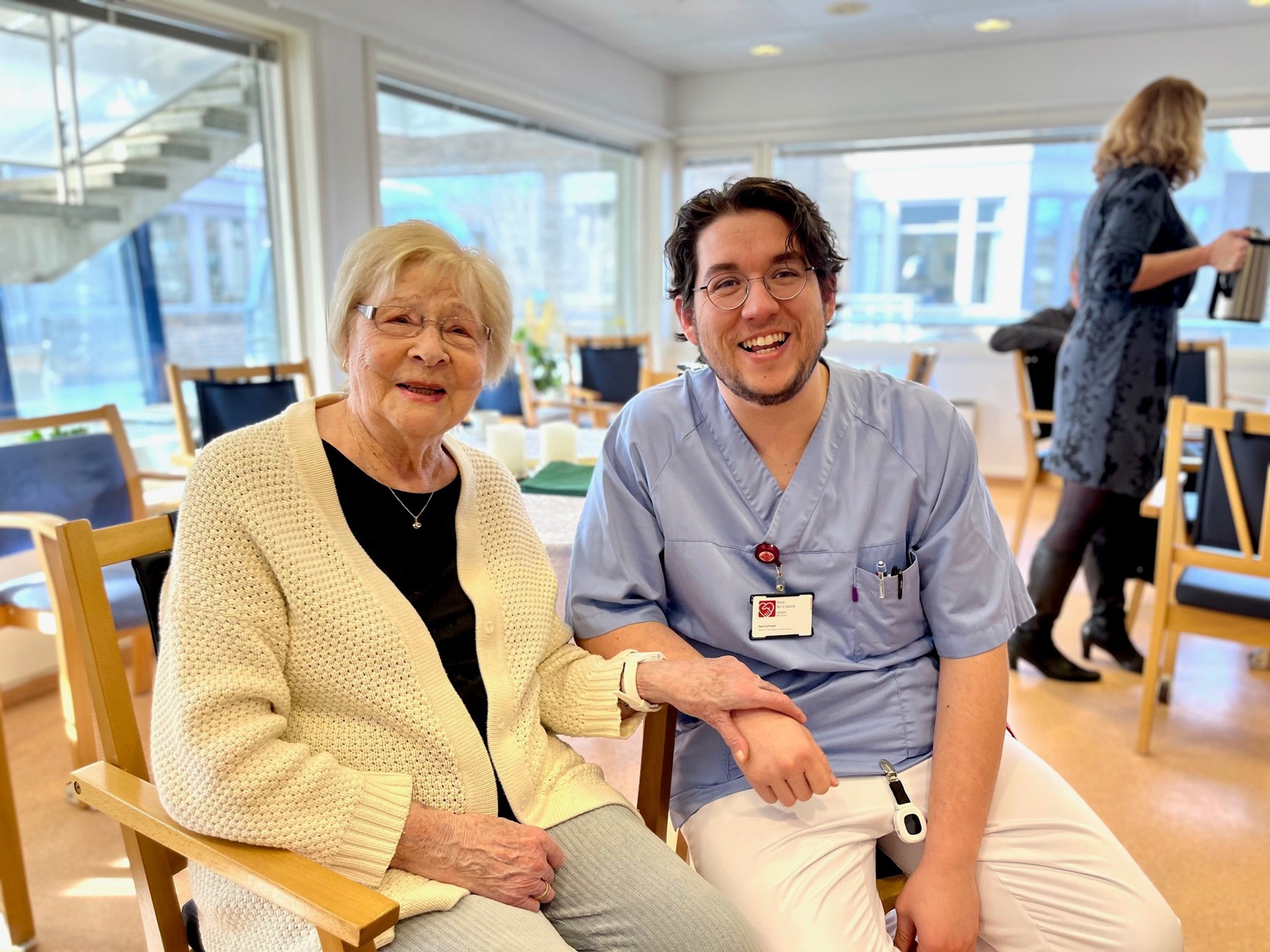 Bildet viser en pasient ved sykehjemmet som sitter ved siden av en pleier. De viser frem armbåndet hun har rundt håndleddet med den nye alarmen.