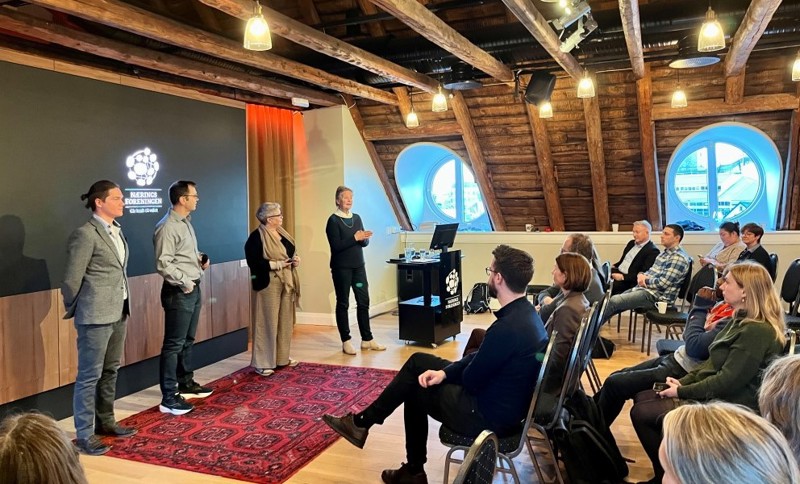 Vi ser et bildet fra møtet, hvor det blir stilt spørsmål fra salen. Vi ser fire personer som står og ser utover salen, hvor det mange folk. 