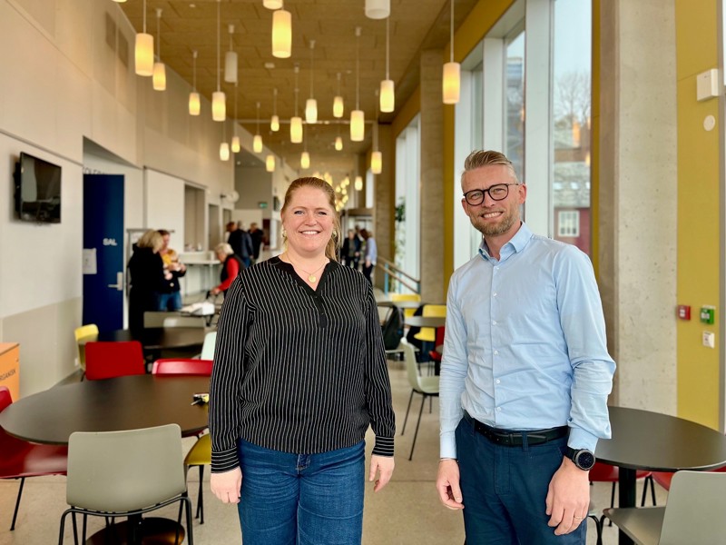 Vi ser Elin Aase og Helge Dragsund som står i foajeen i kulturskolen. Et stort luftig rom, med hengende lamper og bord i bakgrunnen.
