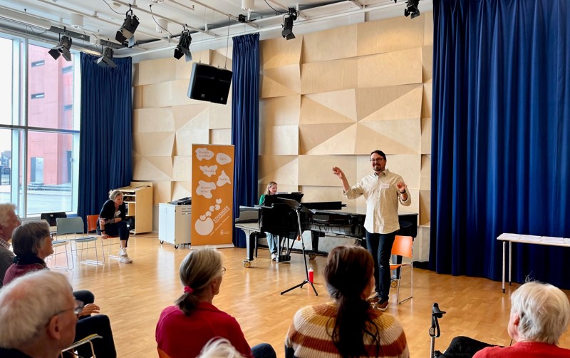 Vi ser dirigenten som står foran på gulvet. Han veiver med armene og viser med hele kroppen hvordan de kan synge. Foran ham sitter det mange kormedlemmer og ser på. 