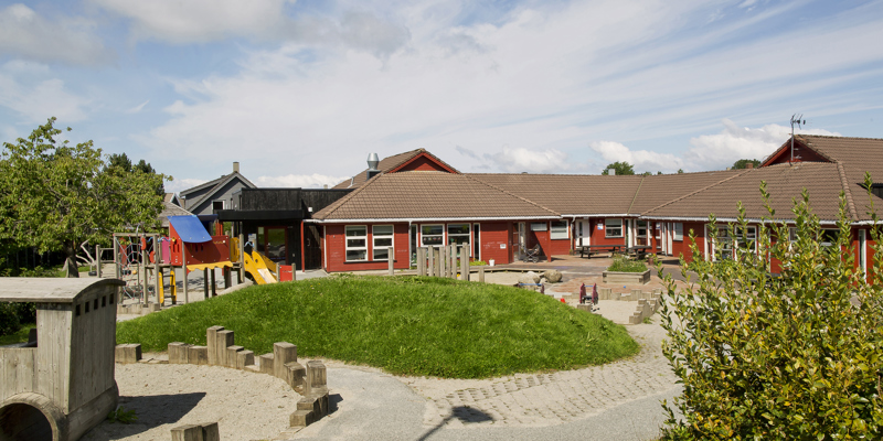 Bildet viser Gullfaks barnehage en sommerdag. Vi ser bygningen og litt av lekeplassen.