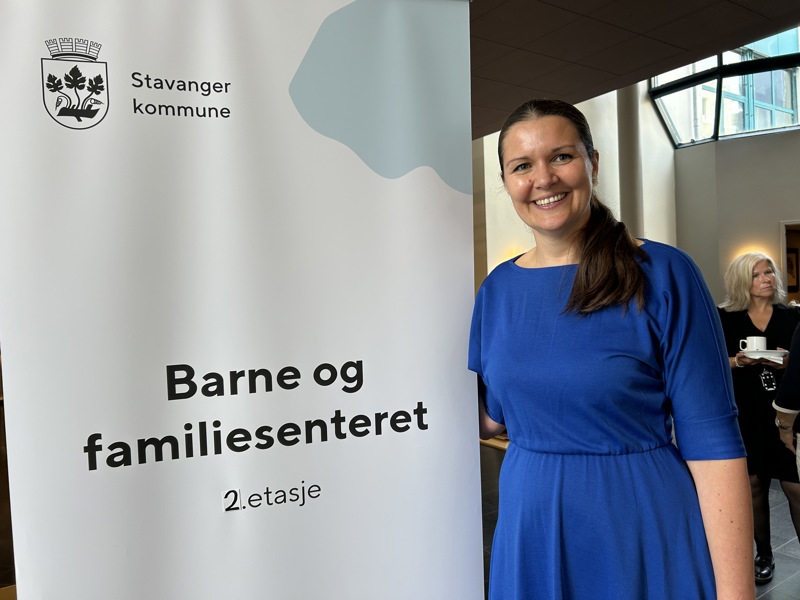 Ingvild Rønneberg Hidle står ved siden av et skilt med barne- og familiesenteret 2. etasje på. Hun har brukt hår, kongeblå kjole og et stort smil.
